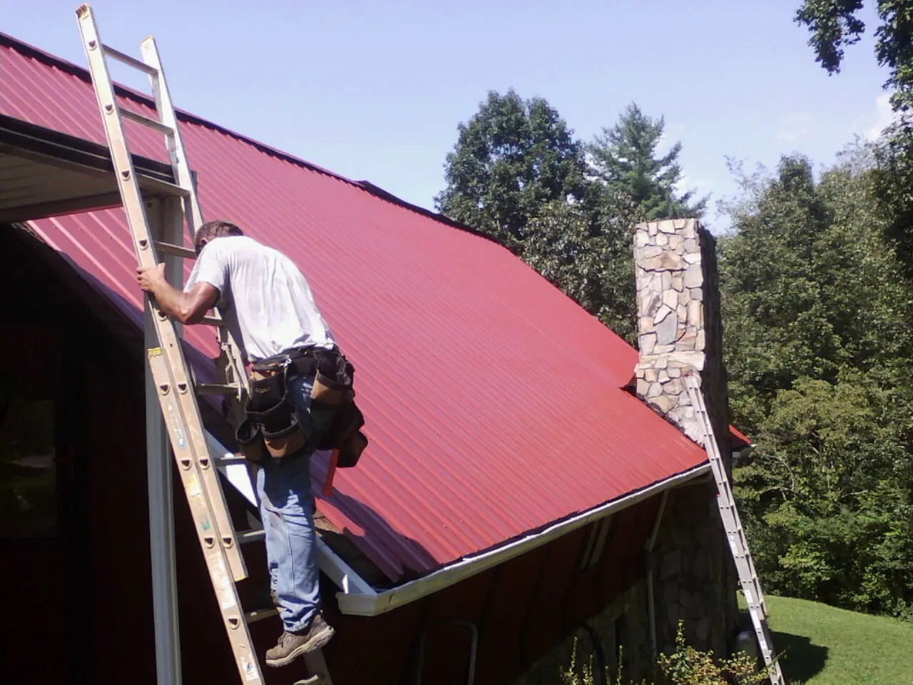 Roofer on a ladder beside a red metal roof and stone chimney — Soffit & Fascia Repair by Roofing Brothers in Applegate
